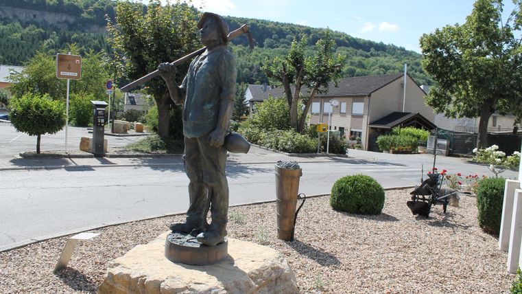 Eine Statue eines Mannes mit einem Pickel steht an einer Straßenecke. Im Hintergrund sind Bäume und Häuser zu sehen.