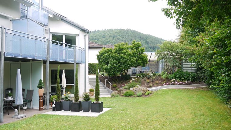 Ein gepflegter Garten hinter einem modernen Haus mit einer grünen Rasenfläche. Es gibt Pflanzen und einen Weg, der zu einer kleinen Terrasse führt.