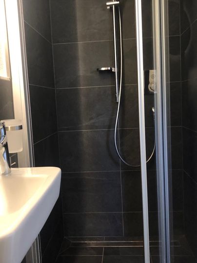 Ein modernes Badezimmer mit einer Glasdusche und einem weißen Waschbecken. Die Wände sind in dunklem Fliesenlook gehalten.