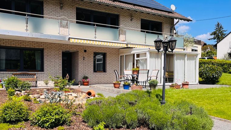 Ein schönes Haus mit Balkon und einem gepflegten Garten. Im Vordergrund sind Blumenbeete und ein gemütlicher Sitzbereich zu sehen.