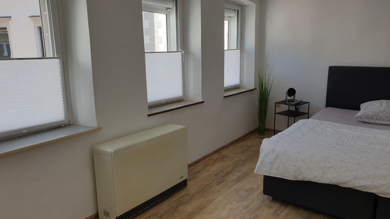 Ein helles Schlafzimmer mit einem modernen Bett und zwei Fenstern. Am Boden liegt ein heller Bodenbelag und eine kleine Pflanze steht neben dem Nachttisch.