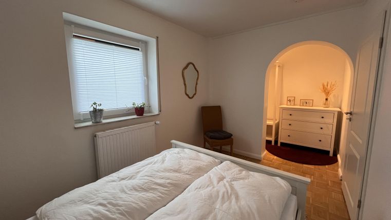 Ein gemütliches Schlafzimmer mit einem Doppelbett und einem Fenster. Im Hintergrund führt ein bogenförmiger Durchgang zu einem weiteren Raum.