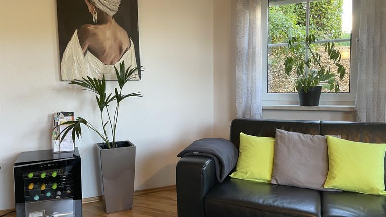 Ein gemütliches Wohnzimmer mit einem schwarzen Sofa und bunten Kissen. An der Wand hängt ein Gemälde und das Fenster bietet Blick auf Pflanzen im Freien.