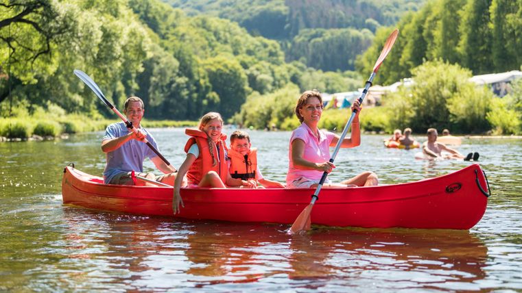 Eine Familie paddelt in einem roten Kanu auf einem ruhigen Fluss. Im Hintergrund sind grüne Bäume und andere Personen, die sich im Wasser entspannen.