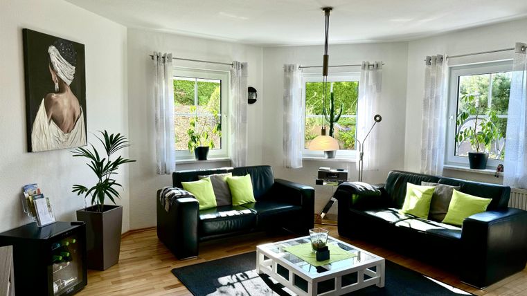 Ein modernes Wohnzimmer mit zwei schwarzen Sofas und grünen Kissen. Große Fenster sorgen für viel Tageslicht und eine angenehme Atmosphäre.