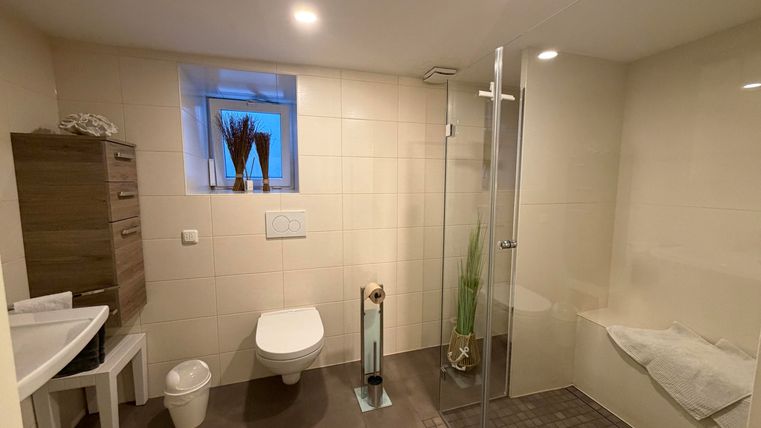 Ein modernes Badezimmer mit einer Duschkabine und einem hängenden WC. Die Wände sind hell gefliest, und es gibt eine große Fensteröffnung für Tageslicht.