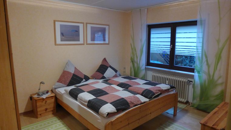 Ein gemütliches Schlafzimmer mit einem Doppelbett und karierten Bettdecken. Licht fällt durch das Fenster und es gibt einige dekorative Pflanzenmotive an der Wand.