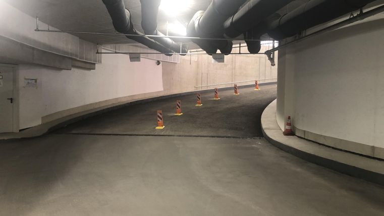 Eine Kurve in einem Parkhaus mit orangefarbenen Absperrungen. Deckenrohre sind sichtbar und das Licht ist gedämpft.