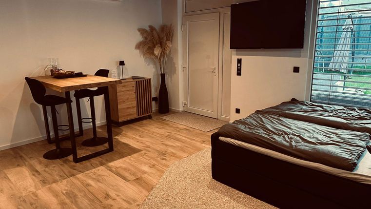 Ein modernes Zimmer mit einem Doppelbett, einem Holzstammtisch und zwei Stühlen. Es gibt große Fenster, die viel Licht hereinlassen.