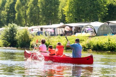 A family is paddling in a red canoe on calm waters. In the background, campsites are visible under trees.