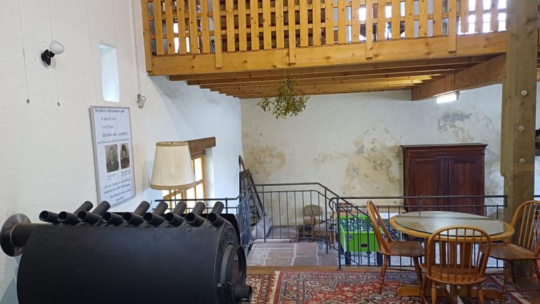 Ein gemütlicher Raum mit einem rustikalen Holzobergeschoss. Im Vordergrund steht ein runder Tisch mit Stühlen und einer antiken Vitrine.