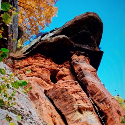 Eine beeindruckende Felsformation mit roten Gesteinen und herbstlichen Bäumen im Hintergrund. Der klar blaue Himmel vervollständigt die Szene.