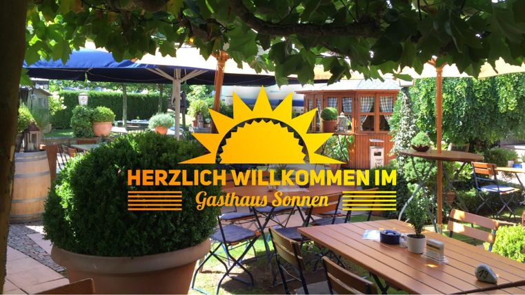 Ein einladender Biergarten mit Tischen und Stühlen unter schattigen Sonnenschirmen. Die Umgebung ist grün und freundlich gestaltet.
