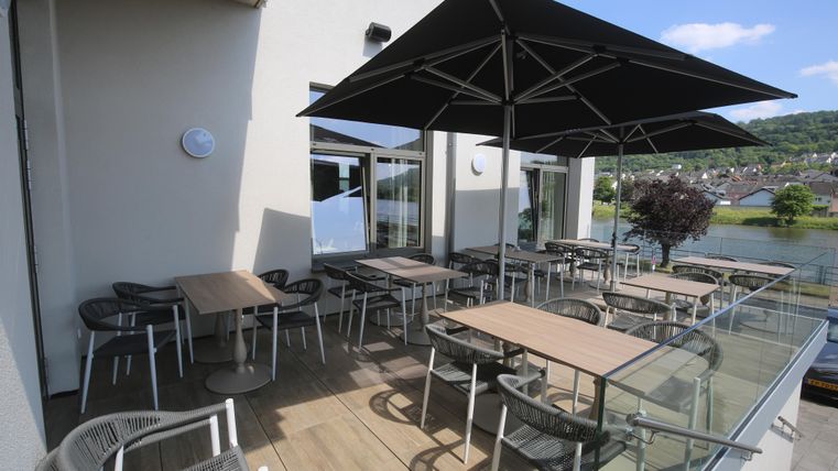 Eine gemütliche Terrasse mit Tischen und Stühlen ist überdacht von einem großen Sonnenschirm. Im Hintergrund ist eine ruhige Wasserlandschaft zu sehen.