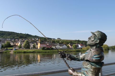 Eine bronzene Statue eines Anglers, der am Fluss steht und angelt. Im Hintergrund sind malerische Häuser und sanfte Hügel zu sehen.