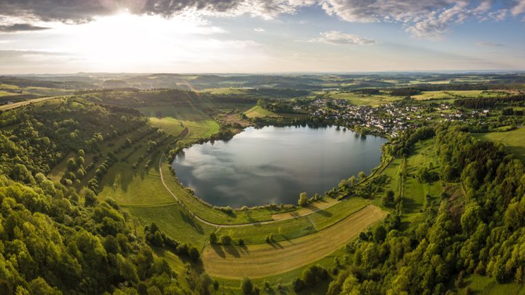 Luftaufnahme des Schalkenmehrener Maars in der Eifel, umgeben von grünen Wäldern und Feldern, mit einem Dorf im Hintergrund.