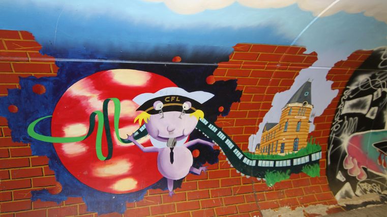 Ein farbenfrohes Wandgemälde zeigt eine cartoonartige Figur mit einer Lokomotive und einer roten Sonne im Hintergrund. Die Szene ist von einer Ziegelsteinwand umgeben.