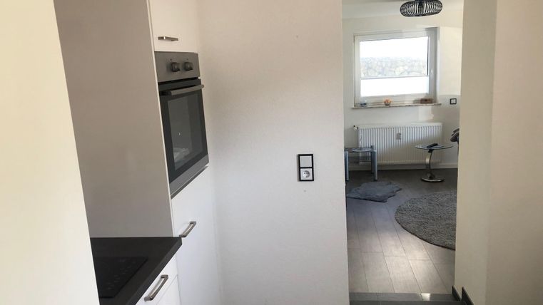 Eine moderne Küche mit einem Backofen und einer hellen Wand. Im Hintergrund ist ein Wohnzimmer mit einem Fenster und einem runden Teppich zu sehen.