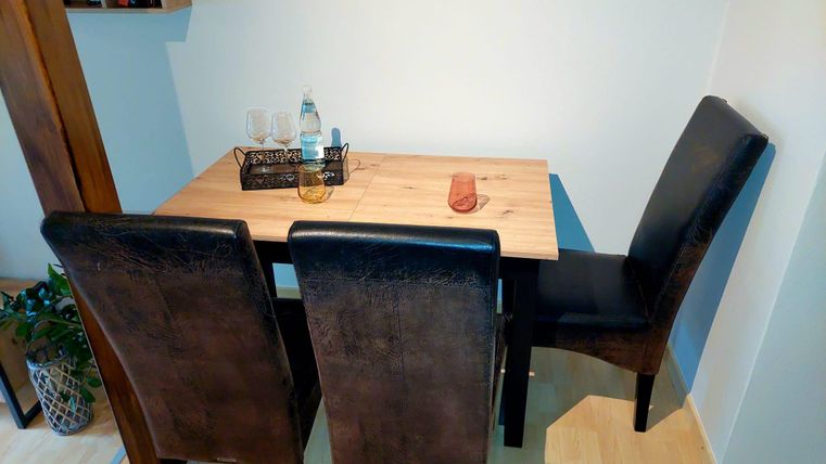 Ein kleiner Esstisch aus Holz mit drei schwarzen Stühlen. Auf dem Tisch befinden sich eine Flasche Wasser und zwei Gläser.