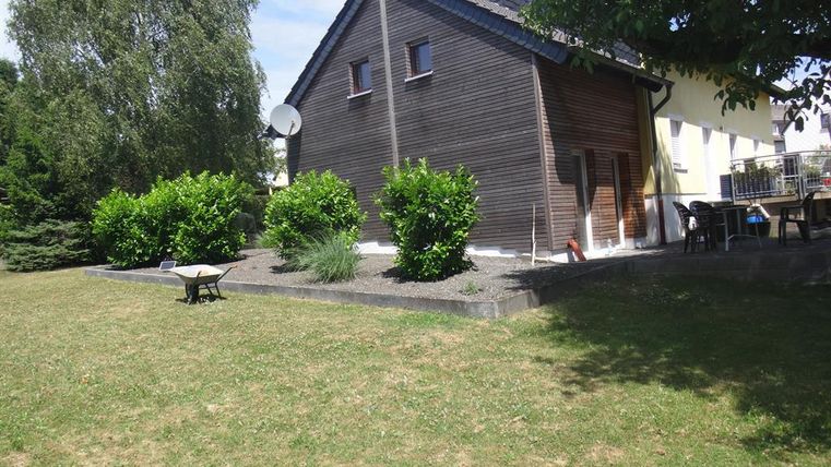 Ein Holzhaus mit einem gepflegten Garten und einigen Bäumen. Die Sonne scheint und der Himmel ist klar.
