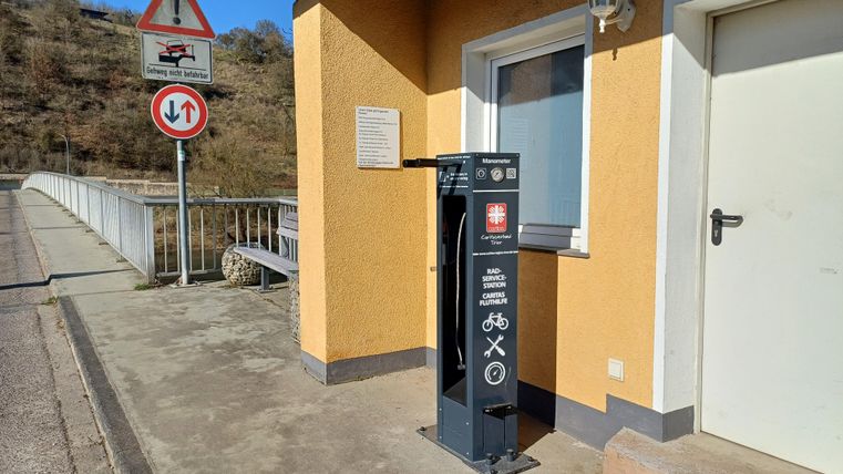 Eine Fahrradservicestation steht vor einem gelben Gebäude mit einem weißen Fenster. Im Hintergrund sind Verkehrszeichen und eine Brücke sichtbar.