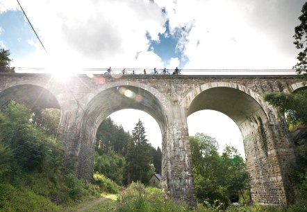 Radfahrer überqueren das Reichensteiner Viadukt der Vennbahn. Die Sonne scheint durch die Wolken, während das Viadukt über eine grüne Landschaft führt., © vennbahn.eu