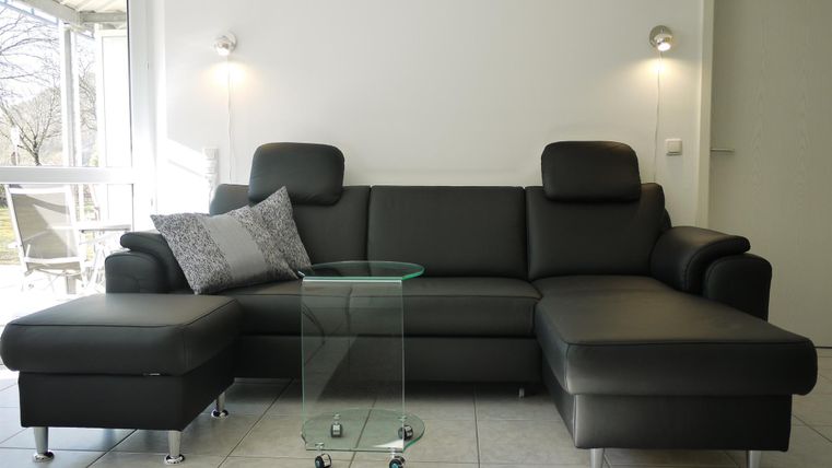 Eine moderne schwarze Couch mit einem zusätzlichen Hocker und einem Glastisch. Der Raum ist hell und freundlich mit großen Fenstern.