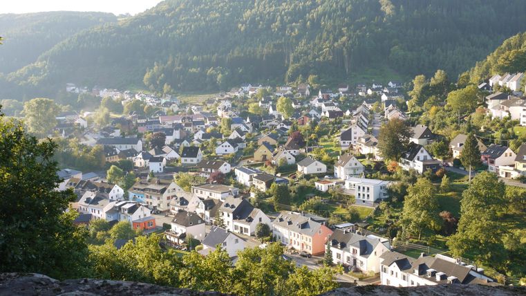 Eine malerische Stadt in einem Tal, umgeben von grünen Hügeln. Die Häuser sind dicht beieinander und das Licht der Abendsonne sorgt für eine warme Atmosphäre.