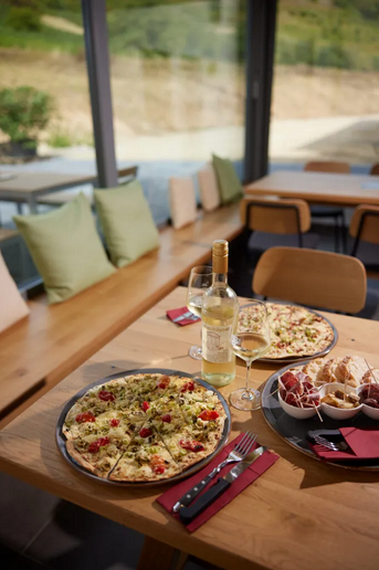 Ein einladender Tisch mit zwei Pizzen, einem Glas Weißwein und einer Auswahl an Antipasti. Große Fenster bieten einen Blick auf die umliegende Natur.