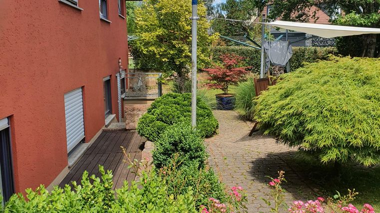 Ein gepflegter Garten mit bunten Blumen und einem schmalen Weg. Im Hintergrund ist ein rotes Haus und ein blauer Himmel mit einigen Wolken sichtbar.