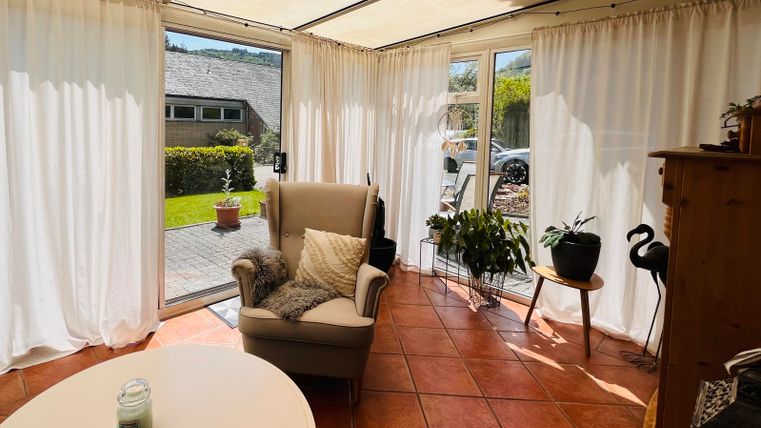 Ein heller Wintergarten mit einem gemütlichen Sessel und einem kleinen Tisch. Durch die Fenster ist der Garten sichtbar, der mit Pflanzen dekoriert ist.