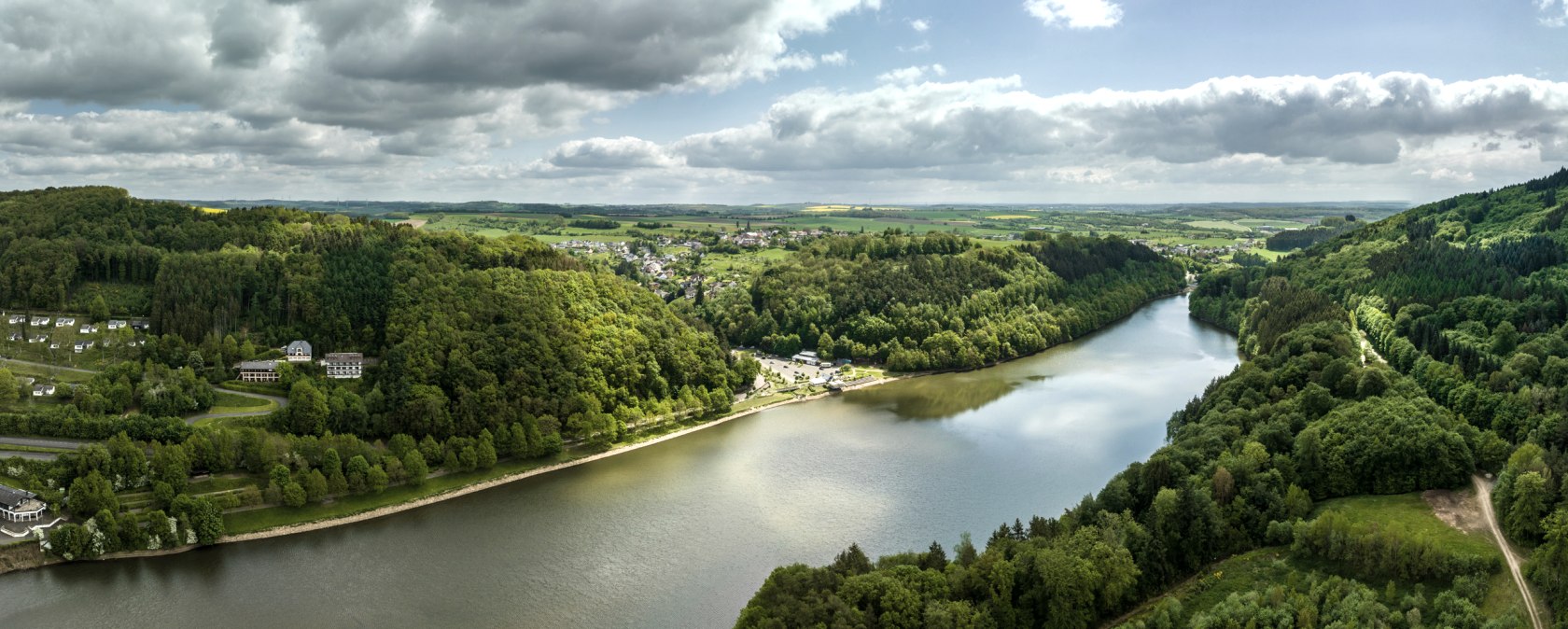 Vue aérienne du lac de barrage de Bitburg à Biersdorf am See, entouré de forêts et de collines verdoyantes. Le ciel est en partie nuageux et en partie ensoleillé., © Eifel-Tourismus GmbH, Dominik Ketz