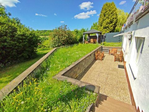 Ein schöner Garten mit grünem Gras und bunten Blumen. Im Hintergrund sind eine Terrasse und ein Pavillon zu sehen.