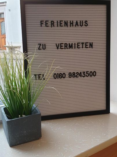 Ein Schild mit der Aufschrift „Ferienhaus zu vermieten“ steht auf einem Tisch. Neben dem Schild befindet sich eine kleine Pflanze in einem Topf.