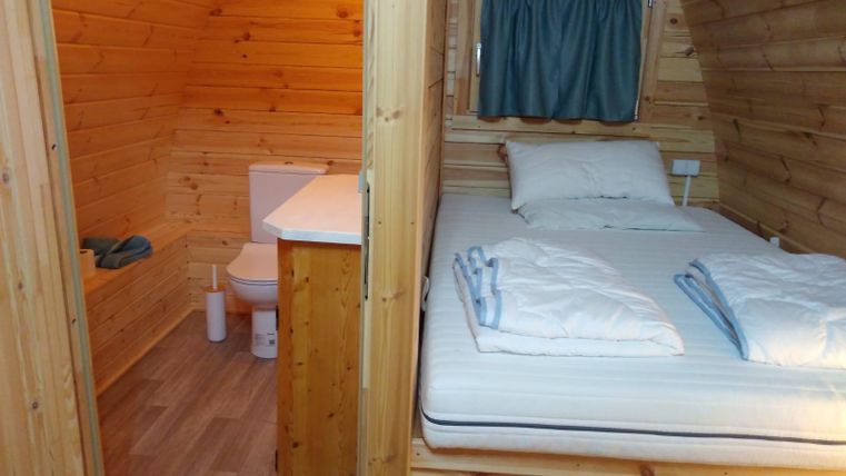 Ein gemütlicher Raum mit Holzvertäfelung, einem Bett und einem kleinen Badbereich. Die Fenster sind mit einem Vorhang verhängt und der Boden ist hell.