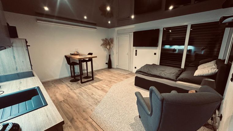 Ein modernes Wohnzimmer mit Holzfußboden und stilvollen Möbeln. Es gibt einen Esstisch, einen Schlafbereich und große Fenster mit Jalousien.