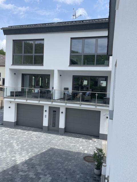 Ein modernes Haus mit großen Fenstern und einem Balkon. Vor dem Haus befindet sich ein gepflasterter Parkplatz mit zwei Garagen.