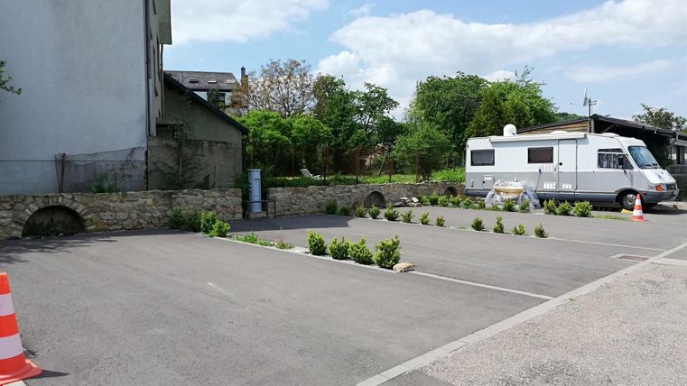 Ein Parkplatz mit einigen kleinen Grünanlagen und einem Wohnmobil. Im Hintergrund sind Bäume und ein Gebäude sichtbar.