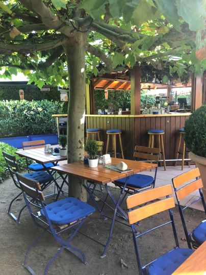 Ein gemütlicher Außenbereich mit Holztischen und Stühlen unter einem Baum. Im Hintergrund ist eine Bar mit verschiedenen Sitzplätzen zu sehen.