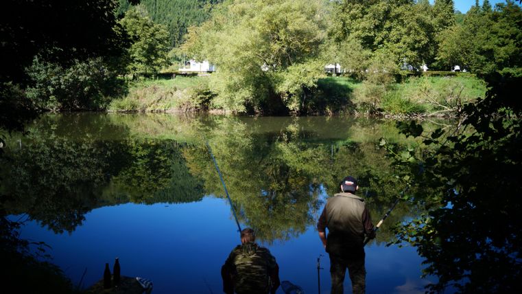 Zwei Angler stehen am Ufer eines ruhigen Flusses, umgeben von grünen Bäumen. Das Wasser spiegelt die Landschaft und den blauen Himmel wider.