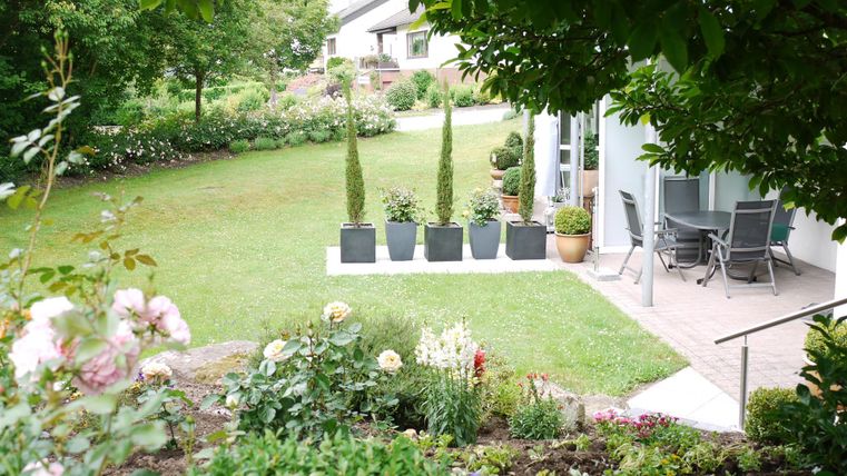 Ein schöner Garten mit gepflegten Blumen und Bäumen. Im Hintergrund steht eine Terrasse mit Möbeln und Kübelpflanzen.