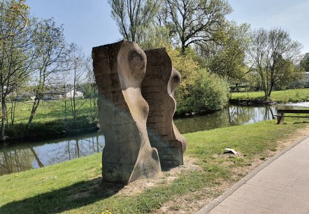 Stenen sculptuur aan de oever van de rivier, omgeven door groen landschap en bomen. Een heldere hemel en een pad zijn ook zichtbaar., © TI Bitburger Land, Steffi Wagner
