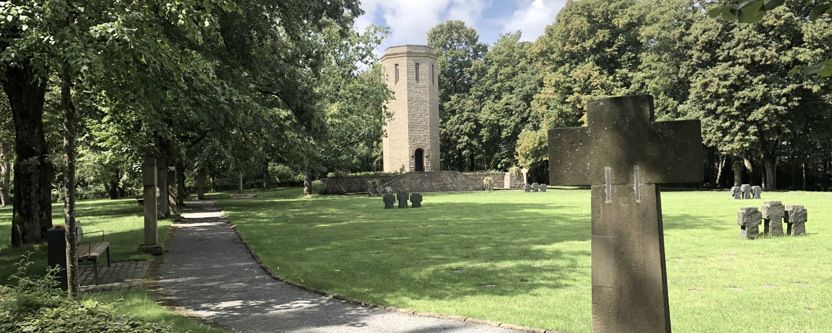 A cemetery with stone crosses, a tower in the background and a path lined with trees. The sky is partly cloudy.