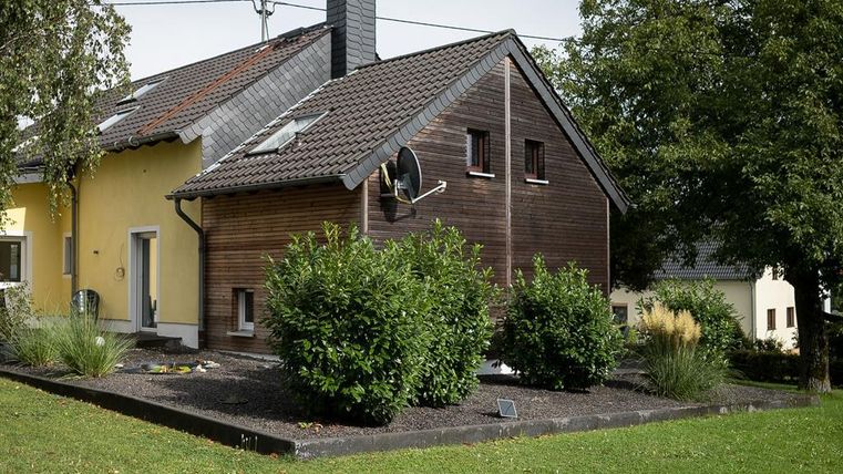 Ein modernes Haus mit einer Holz- und einer gelben Fassade. Der Garten ist mit Sträuchern und Rasen angelegt.