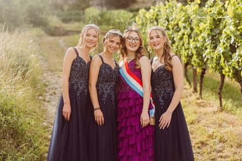 Vier junge Frauen in eleganten Abendkleidern posieren lächelnd in einem Weinberg. Die Atmosphäre ist festlich und fröhlich.