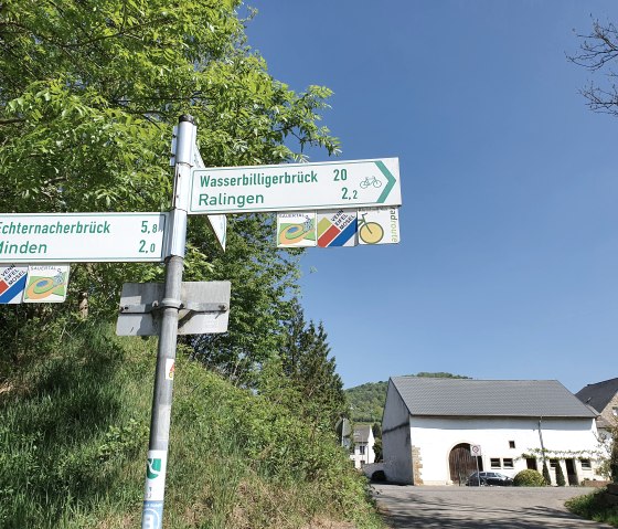 Un panneau indicateur indique les distances jusqu'à Echternacherbrück, Minden, Wasserbilligerbrück et Ralingen. En arrière-plan, un bâtiment, ciel bleu., © Tourist-Information Bitburger Land, Steffi Wagner