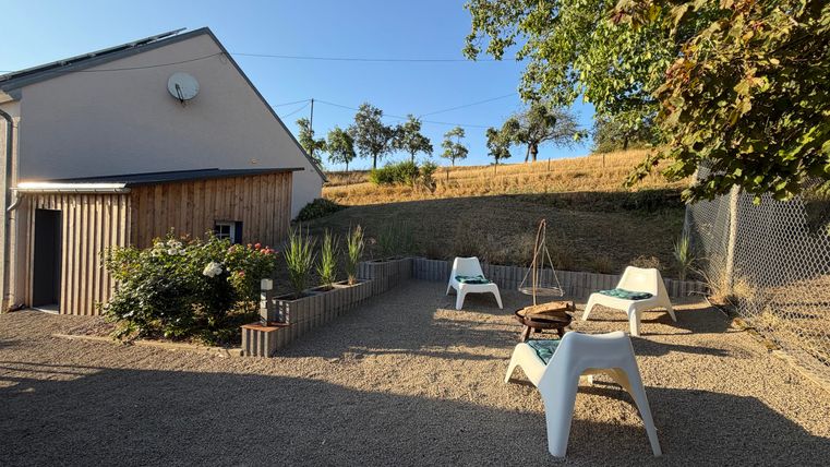 Ein gemütlicher Außenbereich mit einer Terrasse und weißen Stühlen. Im Hintergrund sind Bäume und ein sanfter Hang sichtbar.