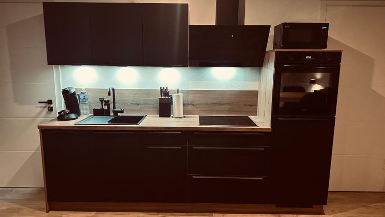 Eine moderne Küche mit dunklen Schränken und einer hellen Arbeitsfläche. Sie ist ausgestattet mit einem Kochfeld, einem Spülbecken und vielen Küchengeräten.