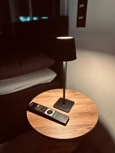 Ein Nachttisch mit einer Stehlampe und einer Fernbedienung. Im Hintergrund ist ein Bett sichtbar.