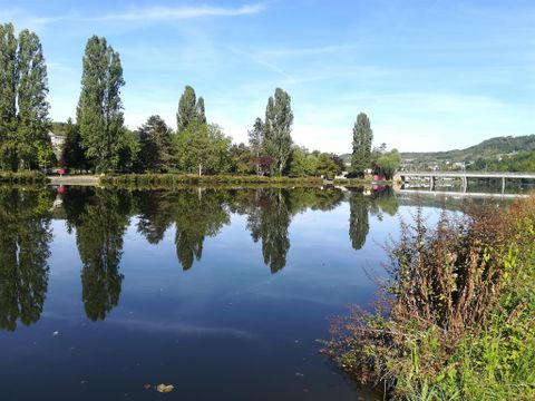 Eine ruhige Wasserfläche mit Spiegelungen von Bäumen. Im Hintergrund sind Brücken und eine grüne Landschaft zu sehen.